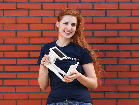 Woman wearing a PATS shirt smiling and holding a PATS-C mounting bracket with solar panel in front of a brick wall
