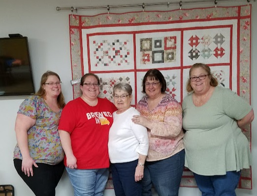 Five woman standing with a quilt on the wall behind them
