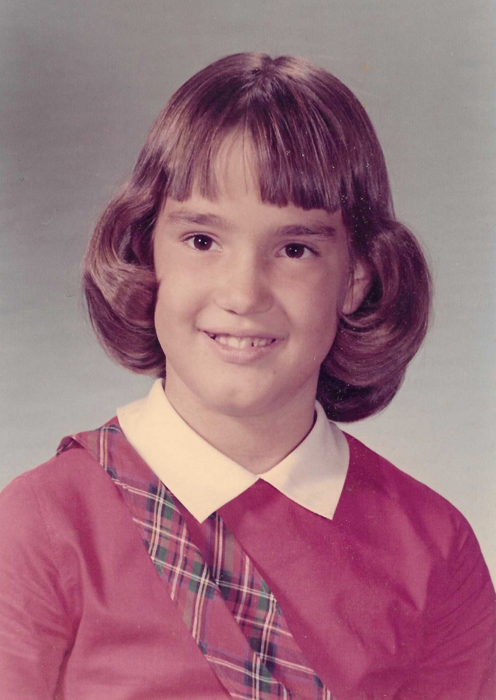 1970 school picture of a brown-haired girl in a red dress with a white pointed collar and red and green plaid diagonal sash