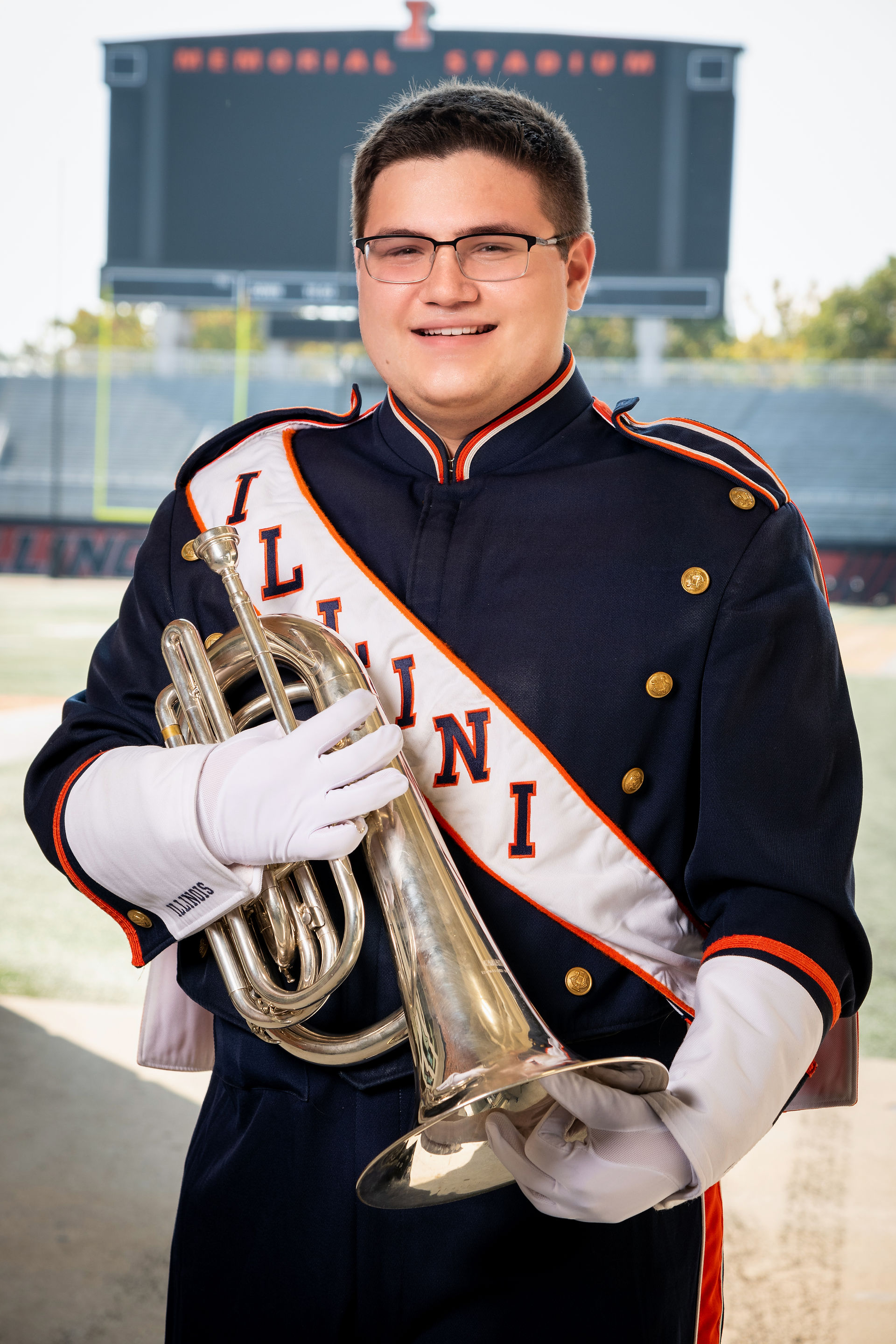 Young man, dressed in a navy blue marching band uniform with a white sash across the chest, is holding a baritone instrument in front of him.