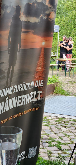Roll-up des Männergruppen-Netzwerks im Vordergrund, während sich drei Teilnehmer des Männerfestivals Chemnitz 2026 im Hintergrund umarmen.