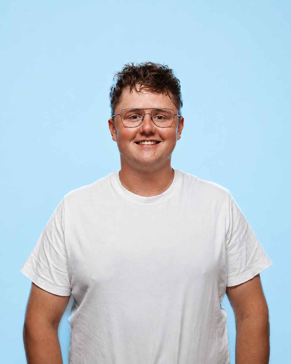 Studio portrait of a smiling young person with curly hair and glasses. The subject is wearing a plain white t-shirt centered against a light blue backdrop.