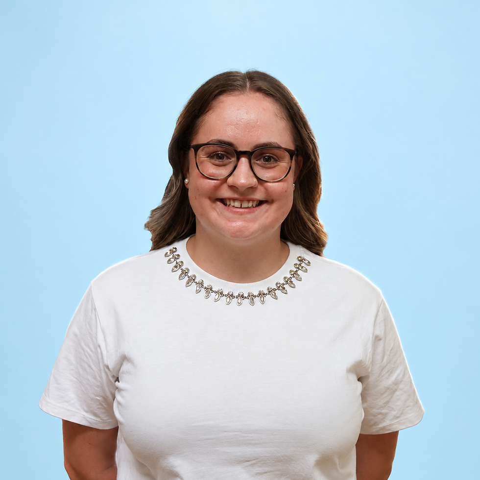 Studio portrait of a young woman with brown wavy hair and glasses, wearing a white t-shirt. She has a friendly, approachable expression against a light blue, monochromatic background.