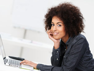 Smiling person with curly hair using a laptop in a bright room, wearing a denim shirt. They rest their chin on a hand, looking content.