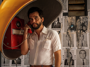 Cena do filme 'O Agente Secreto' com Wagner Moura. O ator está dentro de um orelhão amarelo, falando ao telefone vermelho e olhando seriamente para a frente. Ao fundo, uma parede coberta por cartazes políticos antigos (MDB, ARENA). Logotipo 'Dia a Dica' no canto inferior.