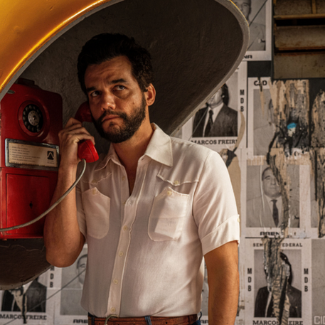 Cena do filme 'O Agente Secreto' com Wagner Moura. O ator está dentro de um orelhão amarelo, falando ao telefone vermelho e olhando seriamente para a frente. Ao fundo, uma parede coberta por cartazes políticos antigos (MDB, ARENA). Logotipo 'Dia a Dica' no canto inferior.