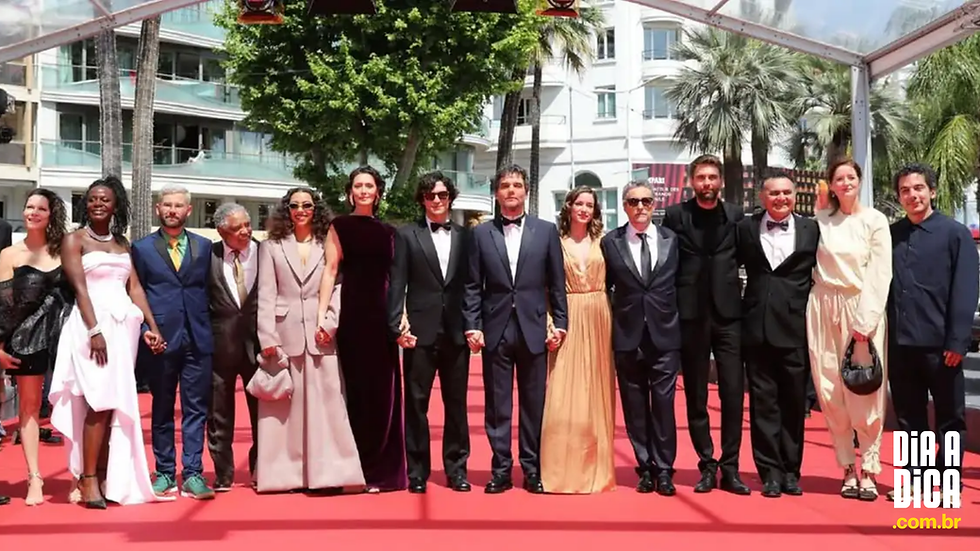 O elenco do filme 'O Agente Secreto' posa de mãos dadas no tapete vermelho do Festival de Cannes. O grupo veste trajes de gala variados sob a luz do dia, com palmeiras e edifícios brancos ao fundo. Logotipo 'Dia a Dica' no canto inferior.