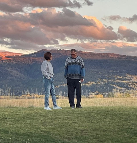 A couple standing outside in the grass with the mountains and clouds in the background