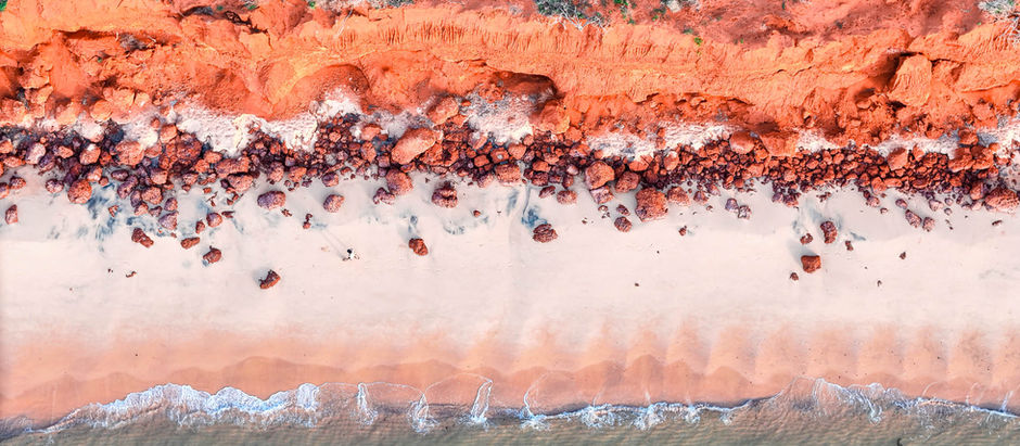 Overhead drone shot of the red sand cliffs meeting white sand beach and calm shallows at Francois Peron National Park, with a lone person walking along the shore for scale.