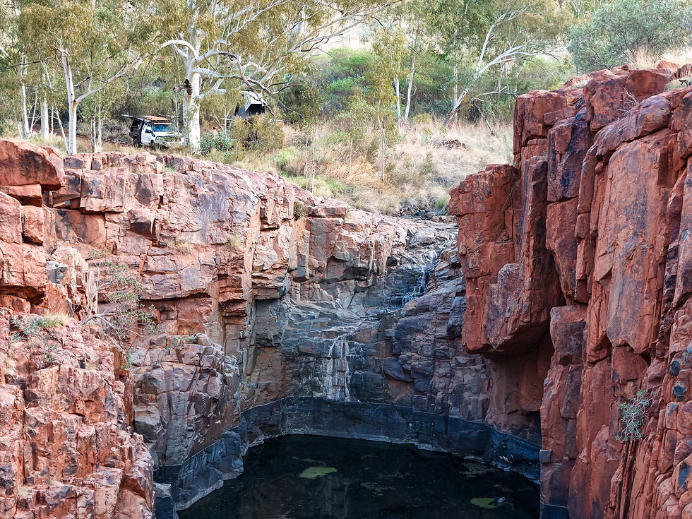Deep waterhole carved into red and grey banded rock at Three Pools near Newman, Western Australia, with a Toyota Hilux 4WD and rooftop tent visible camped on the cliff edge above the gorge among white-trunked river gums. Photograph taken from the rim looking down into the still dark water below.