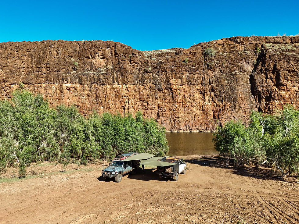 Two 4WDs camped under awnings beside a still river, with a towering layered red rock cliff face rising at Carawine Gorge.