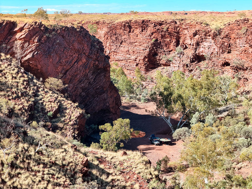 Toyota Hilux 4WD with awning extended camped beside the waterhole at Kalgan's Pool near Newman, surrounded by river gums and steep layered red rock cliff walls. Aerial photograph showing the remote canyon campsite from the clifftop above.