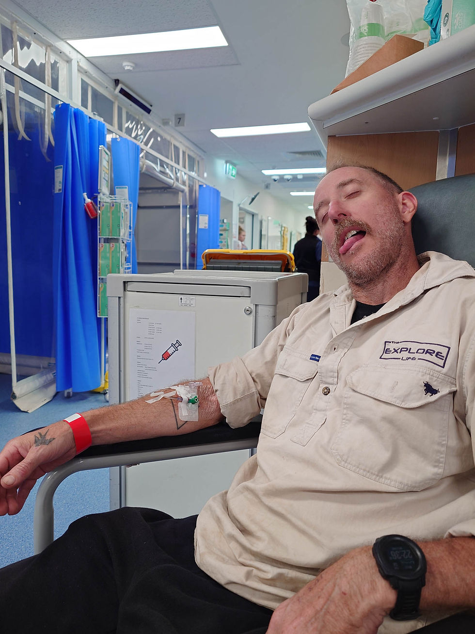 Man with an IV in his arm sits in a hospital hallway, eyes closed and tongue out, wearing beige shirt and watch. Blue curtains in background.