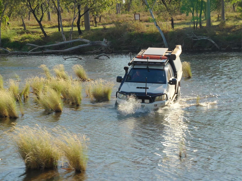 A white four-wheel drive loaded with rooftop gear crossing a wide river, water spraying at the wheels, with grassy banks and gums behind.