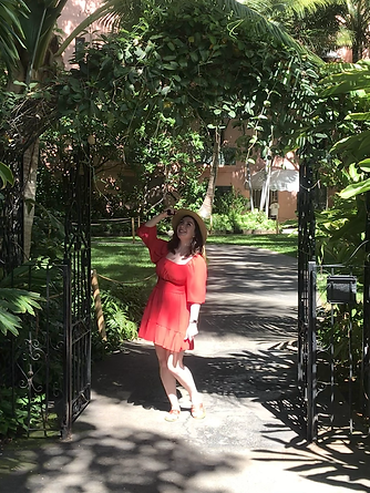 A photo of me, Caoilfhinn, in a red dress smiling while looking up. I'm holding the edge of a floppy hat underneath a black iron garden gate, where a path leads through a garden in the background.