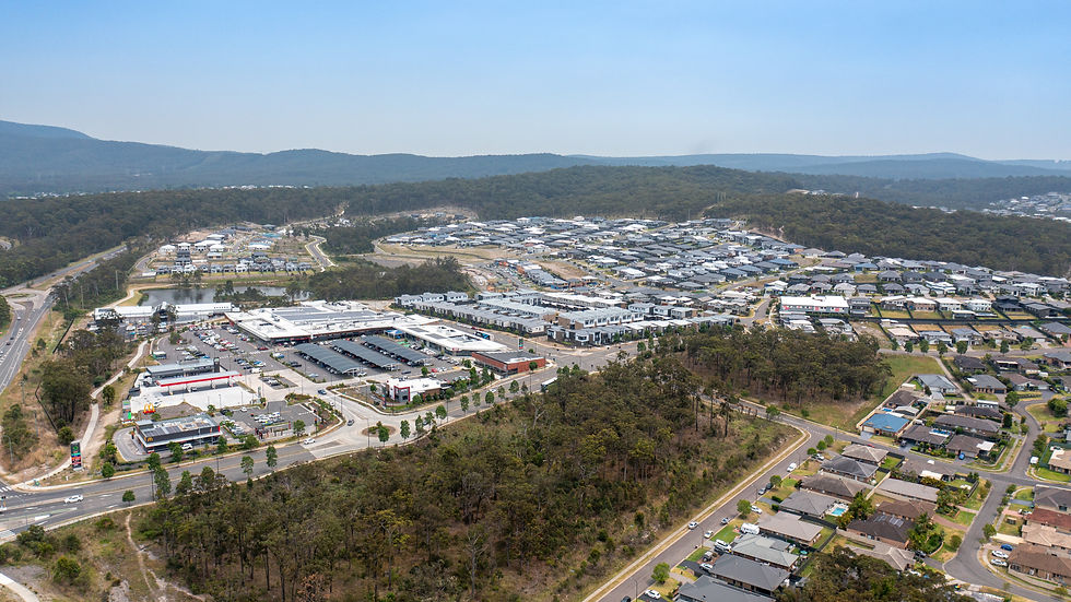 Aerial view of a suburban area with houses, a shopping center, roads, and a forested landscape. Clear sky and distant hills in the background.
