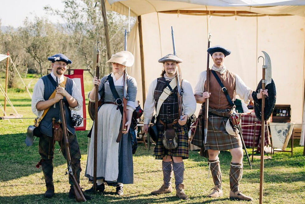 Four people in historical attire hold weapons, standing on grass by a tent. They're smiling, and the scene is bright and sunny.