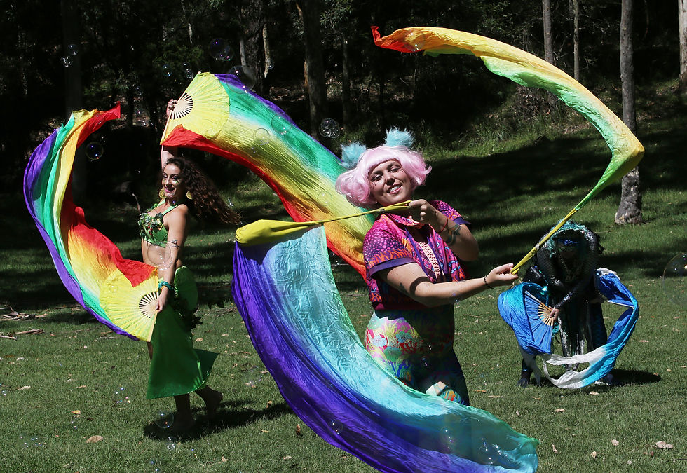 People dance with rainbow ribbons in a sunny park, surrounded by bubbles. Vivid costumes add to the joyful, vibrant scene.