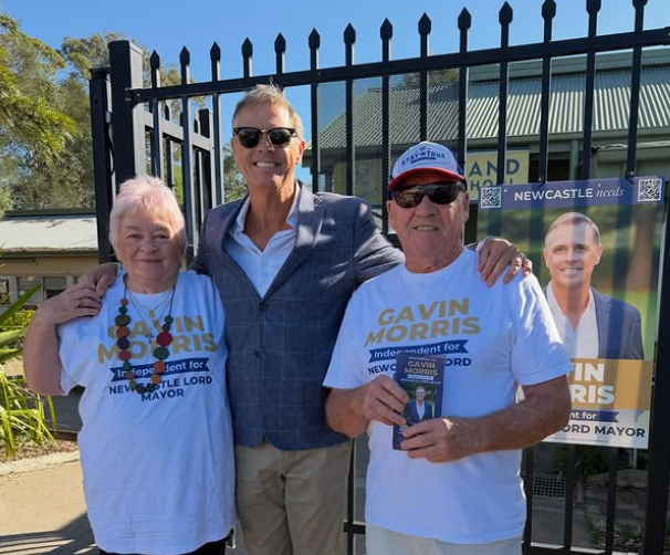 Three people smile in front of a fence with campaign posters. Two wear "Gavin Morris" T-shirts, holding a brochure. Sunny day setting.