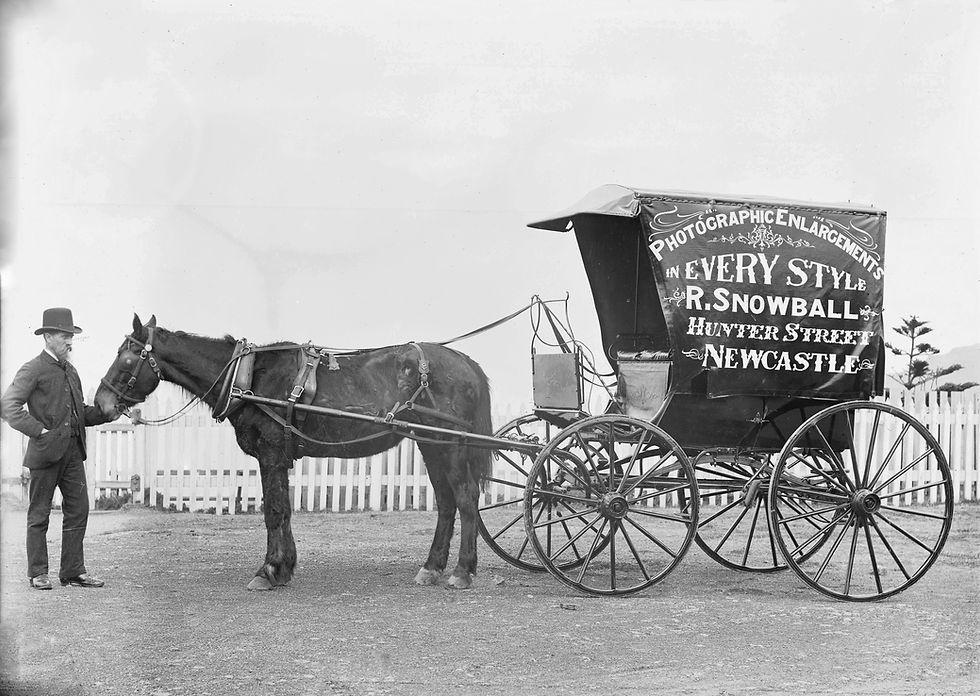 Man stands beside a horse-drawn carriage with a sign reading "Photographic Enlargements, R. Snowball, Hunter Street, Newcastle." White fence behind.