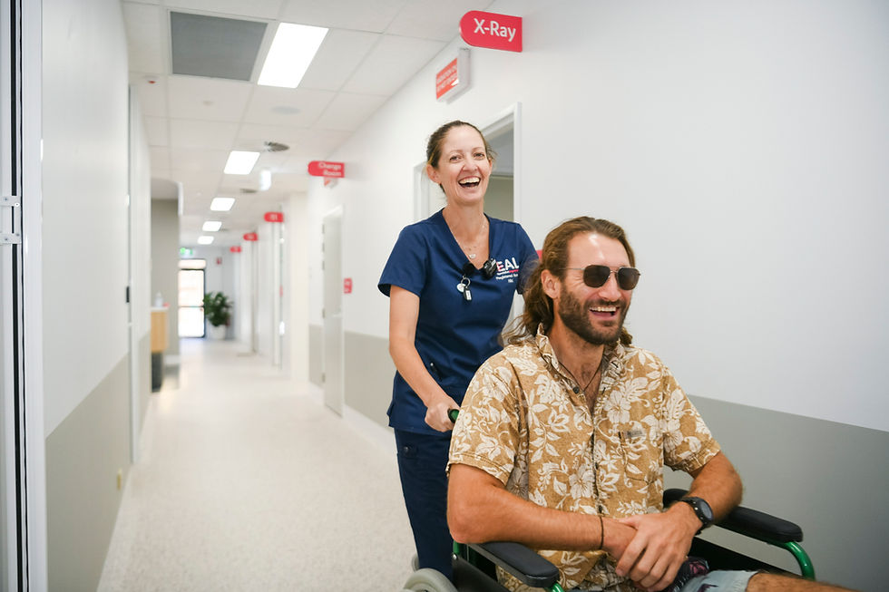 Nurse in blue scrubs happily pushes a smiling man in a wheelchair down a hospital hallway. Signs for X-Ray and Change Room visible.