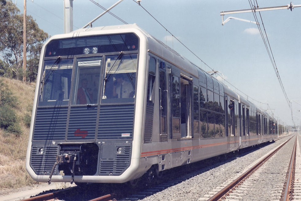 Silver train with red arrow graphic travels on tracks under a clear sky. Trees line the right side. Passengers visible in front cab.