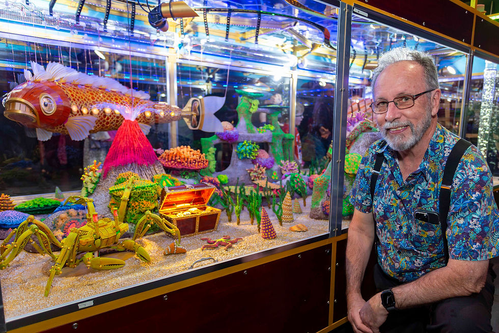Man smiles by a colorful aquarium display featuring a mechanical fish and crab, vibrant coral, and treasure chest. Bright and whimsical.