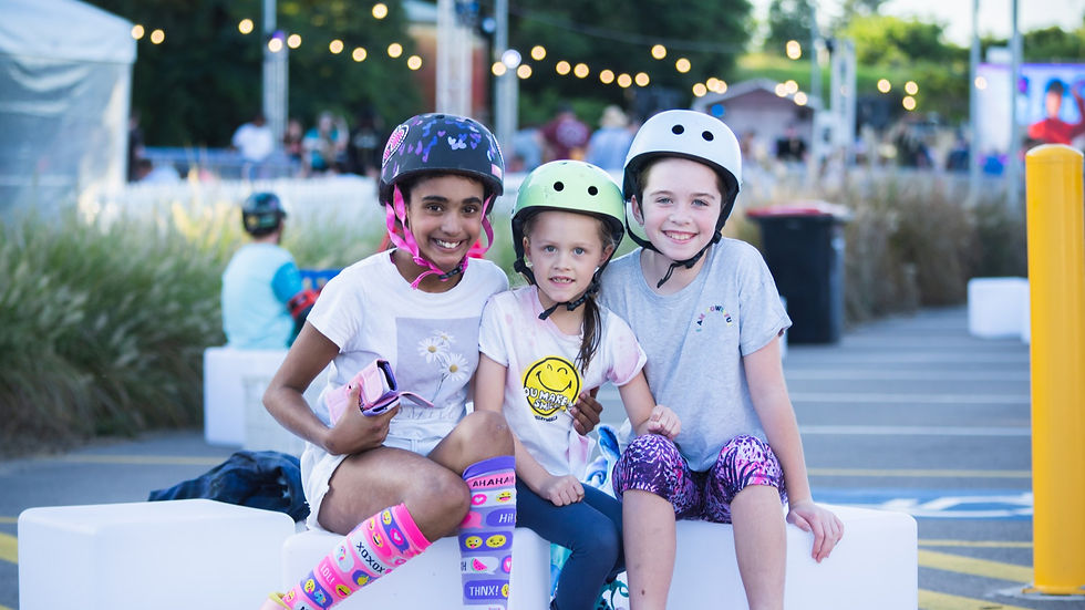Three smiling kids in helmets sit on white blocks outdoors. They wear colorful outfits with emoji and floral patterns. String lights hang above.