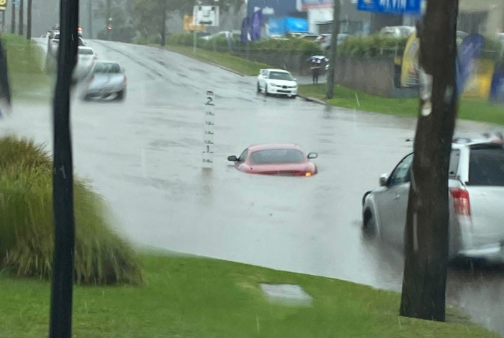 A red car partially submerged in floodwater on a rainy street, surrounded by moving traffic and blurred greenery, evokes a tense mood.