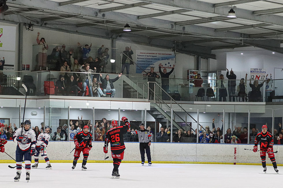 Hockey players in action on ice, with a crowd cheering enthusiastically from the stands. Red and blue uniforms, indoor rink setting.