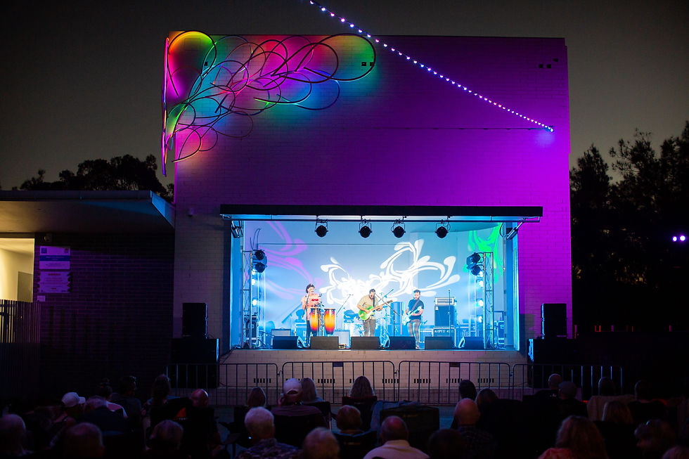 Outdoor concert at night with musicians on stage. Colorful abstract light patterns and spotlights illuminate the scene. Audience seated in front.