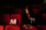 Woman sits in red theater seats, a teddy bear and popcorn beside her. She has a pink ribbon on her jacket, looking pensive in dim light.