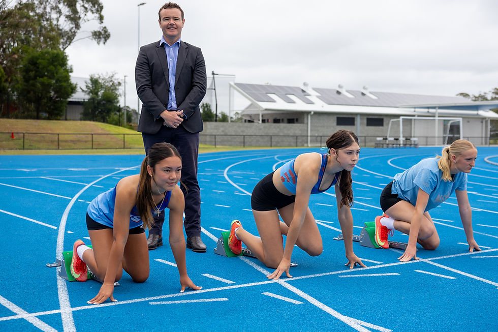 Three female athletes in starting positions on a blue track, with a man in a suit standing behind. Overcast sky and sports facility visible.