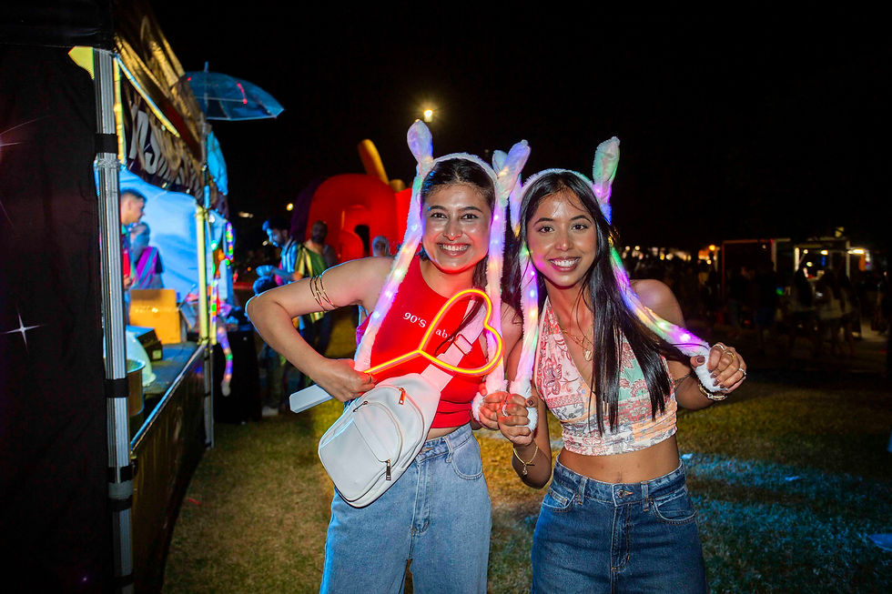 Two women at a nighttime festival wear glowing bunny ear hats, smiling and holding a neon heart. Bright lights and stalls are in the background.