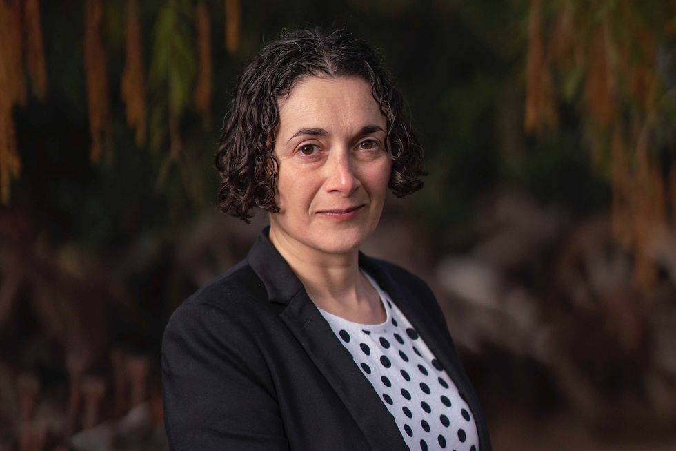 A woman with curly hair in a black blazer and polka dot shirt poses outdoors, with a blurred natural background. She has a calm expression.