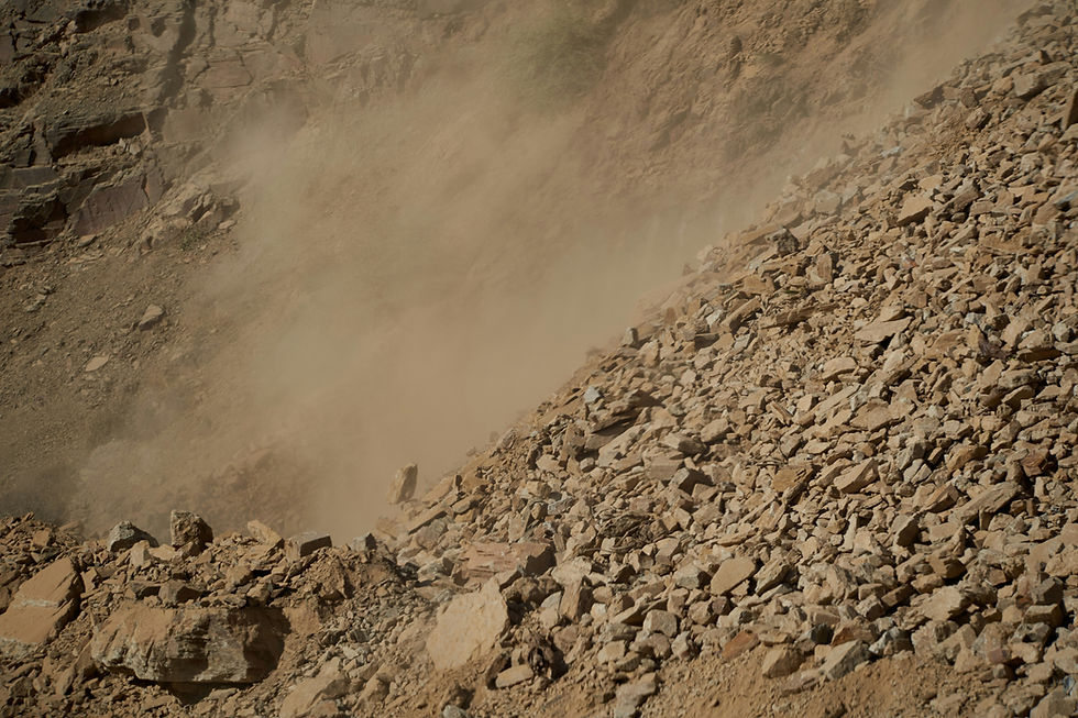 Dust clouds rise over a rocky landslide in a barren canyon, with tan rubble spilling down steep cliff walls.