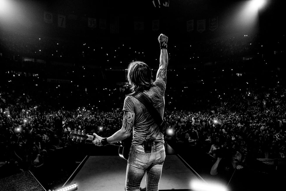 Musician on stage raises a fist, holding a guitar. Energetic crowd with phones lit up surrounds him in a dark arena, creating a lively mood.