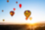 Hot air balloons with colorful patterns float at sunrise over a misty landscape. Text on one balloon reads: Balloon Aloft. Calm mood.