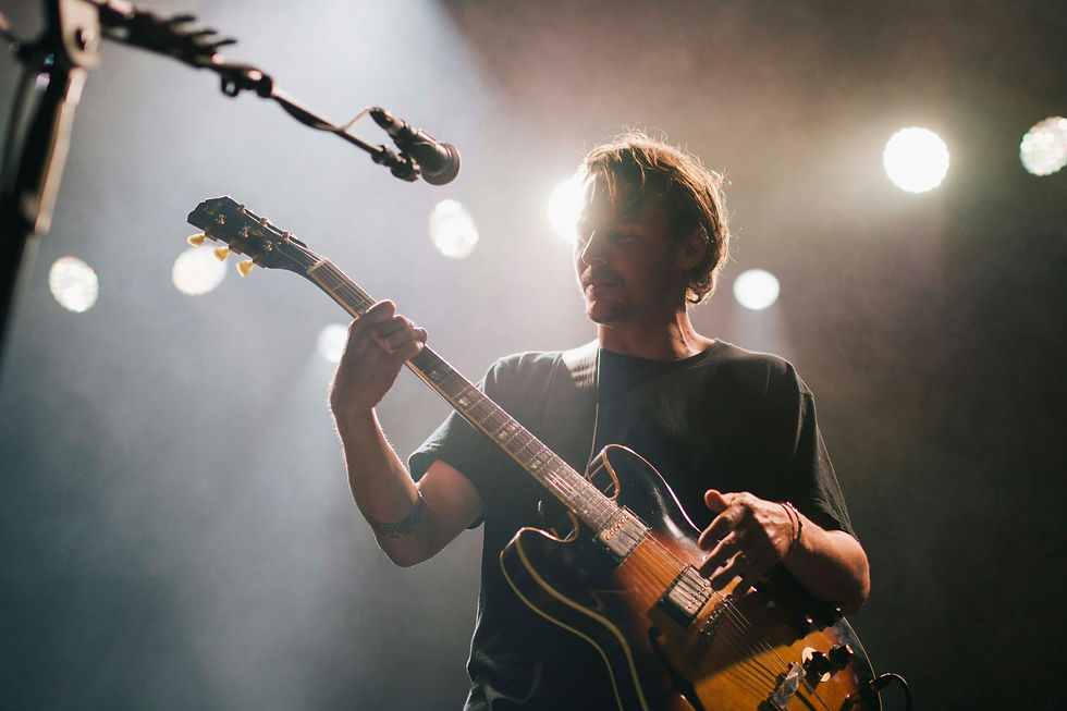 Musician playing guitar on stage, lit by bright spotlights in the background, creating a moody atmosphere. Dark clothing, focused expression.
