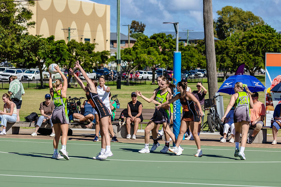 Netball game in progress outdoors with players in green and black uniforms reaching for the ball. Spectators and trees in the background.