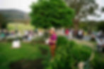Crowd gathered in a lush garden setting for an outdoor event, with a man in a hat holding papers and a drink, surrounded by greenery and sculptures.