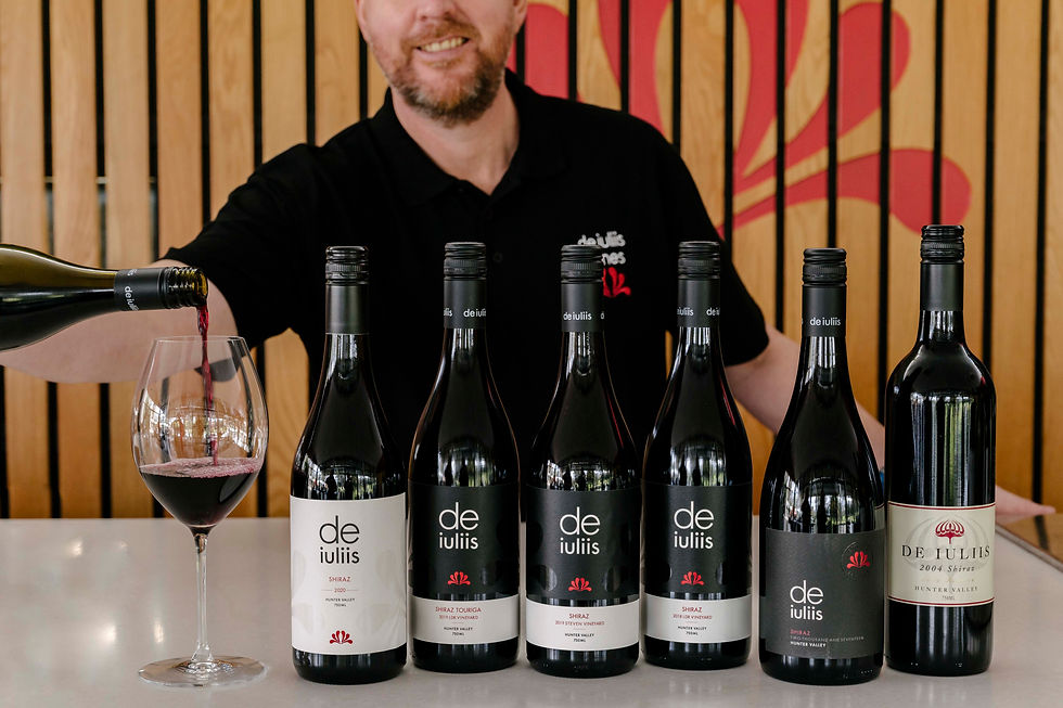 Man pouring red wine into a glass; five de iuliis wine bottles lined up. Warm wooden backdrop with red accents.