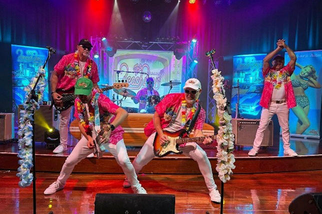 Band in colorful Hawaiian shirts plays guitars energetically on stage with vibrant lights and floral decorations. Background shows beach-themed images.