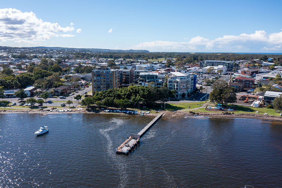 Aerial view of a town by a river, with a pier extending into the water. Buildings, trees, and a boat are visible under a partly cloudy sky.