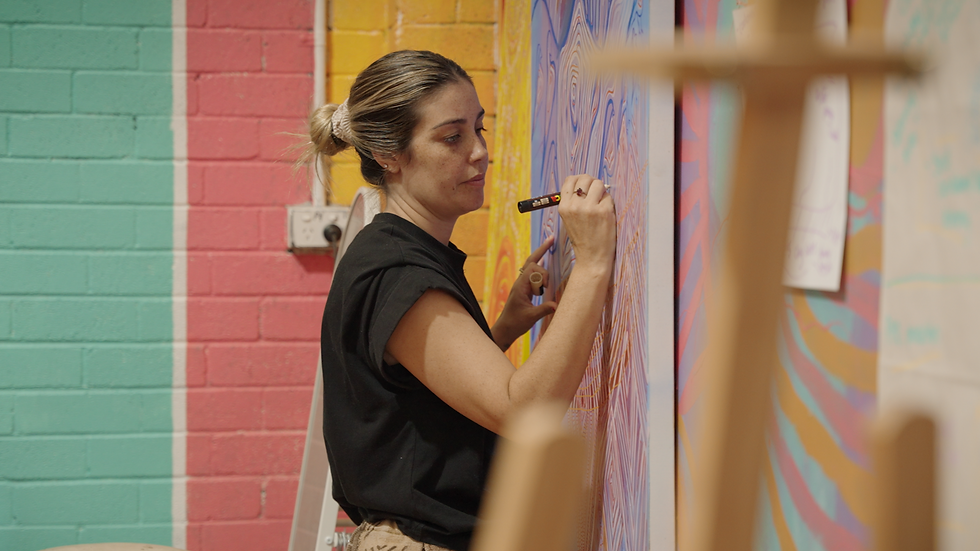 Artist painting colorful swirls on a wall indoors. Background has turquoise, red, and yellow sections. Focused expression with a brush in hand.