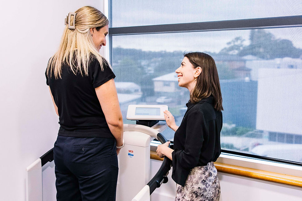 Two women in a bright room are smiling and talking by a window. One gestures to a digital monitor on a medical device.