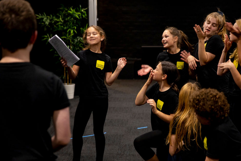 Children in black shirts with yellow logos, one holding a script, act joyfully in a rehearsal space with a potted plant in the background.