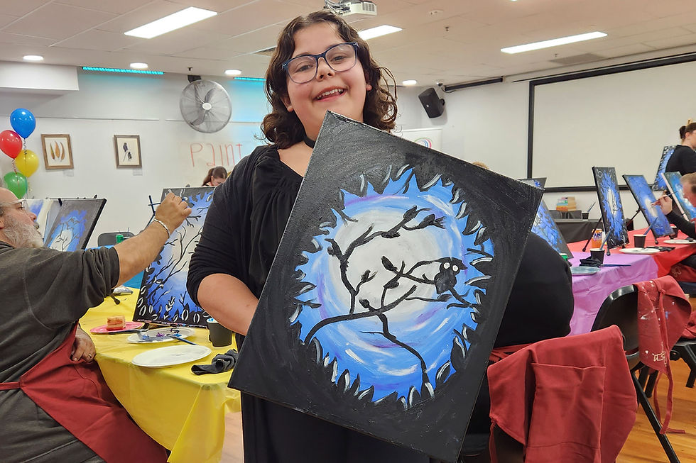 A person smiles, holding a painting of a tree silhouette against a blue and white background. Art class with colorful balloons and tablecloths.