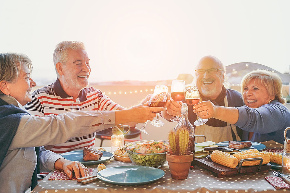 Four smiling older adults toast with wine at a sunny outdoor dinner table with salad, corn, and grilled meat.