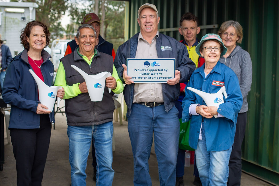 Group of smiling people holding watering cans and a sign about Hunter Water's Love Water Grants. Outdoor setting, casual attire.